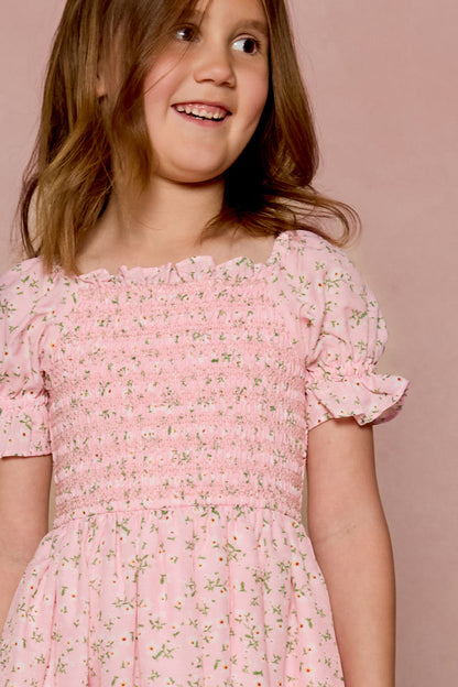 A young girl smiles in the Ivy City Co Mini Valencia Dress in Pink Ditsy Floral, featuring a smocked bodice and short sleeves, as she stands before a matching pink background.