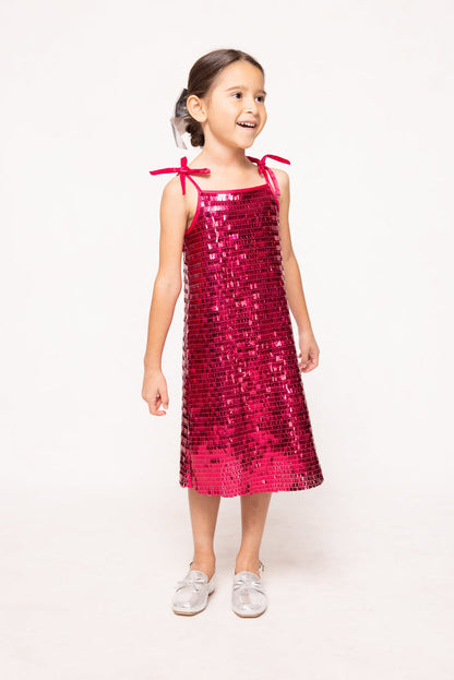 A young girl smiles while wearing the Ivy City Co Mini Zelda Dress in Fuchsia Shingle Sequin, featuring shoulder ties, and silver shoes, standing against a plain light background.
