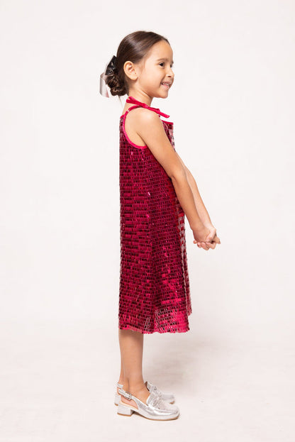 A young girl stands sideways, smiling, dressed in Ivy City Co’s Mini Zelda Dress in Fuchsia Shingle Sequin—a dazzling sleeveless party dress—paired with silver shoes. Her hair is styled in a neat bun and her hands are gracefully clasped in front of her.