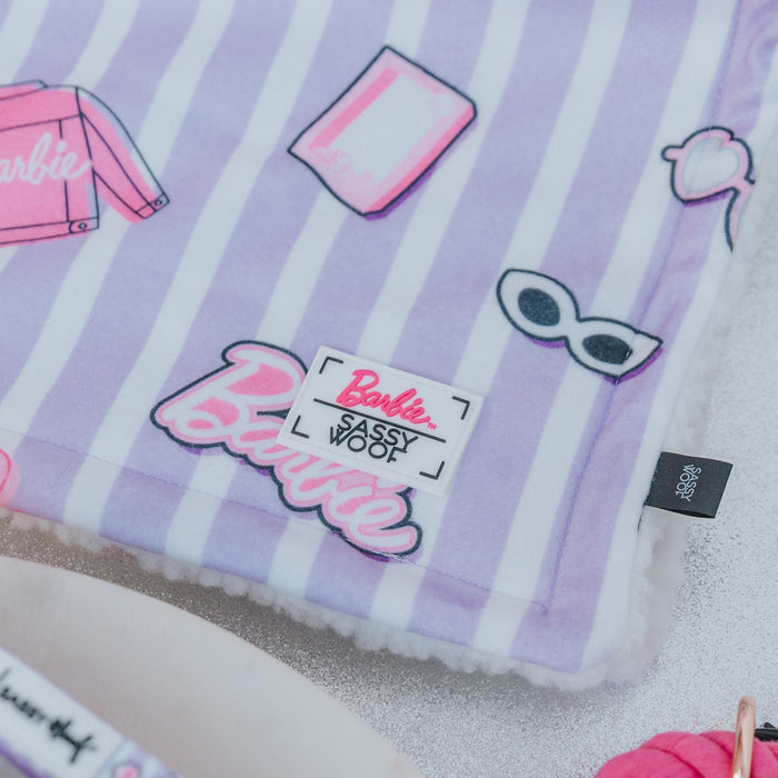 The Sassy Woof Pet Blanket - BARBIE™ Closet Goals is a soft, striped lavender dog blanket with white lines and Barbie-themed illustrations. It features a white patch reading "Barbie x Sassy Woof" and a black tag on the right side.