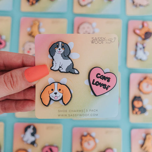 A hand with orange nail polish holds a Sassy Woof Shoe Charm - Cavs card, displaying three vibrant charms: playful dog faces and a heart that reads "Cavs Lover," with other pastel-branded breed accessories blurred in the background.