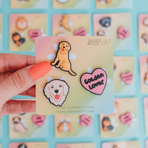 A hand with orange-painted nails holds Sassy Woof's Shoe Charm - Goldens set, featuring a cartoon golden retriever, a retriever face, and a pink "Golden Lover" heart charm, with similar breed accessories in the vibrant background.
