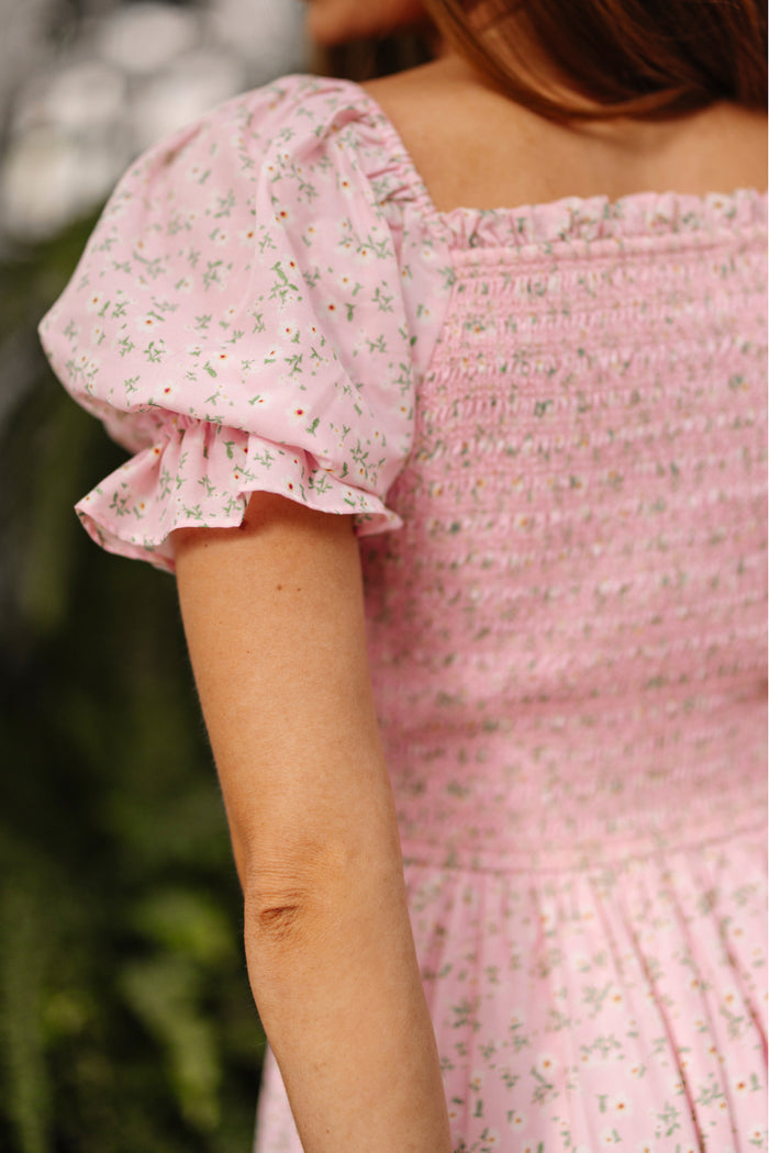 A person is shown from the back wearing Ivy City Co’s Valencia Dress in Pink Ditsy Floral, a maternity-friendly dress with ruffled short sleeves and a shirred bodice, set against softly blurred greenery.