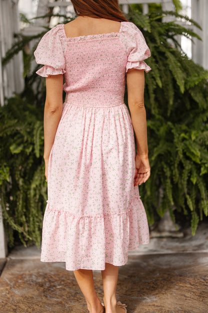 A woman stands among green ferns on a stone floor, wearing the Ivy City Co Valencia Dress in Pink Ditsy Floral, featuring a smocked, maternity-friendly fit and ruffled hem.
