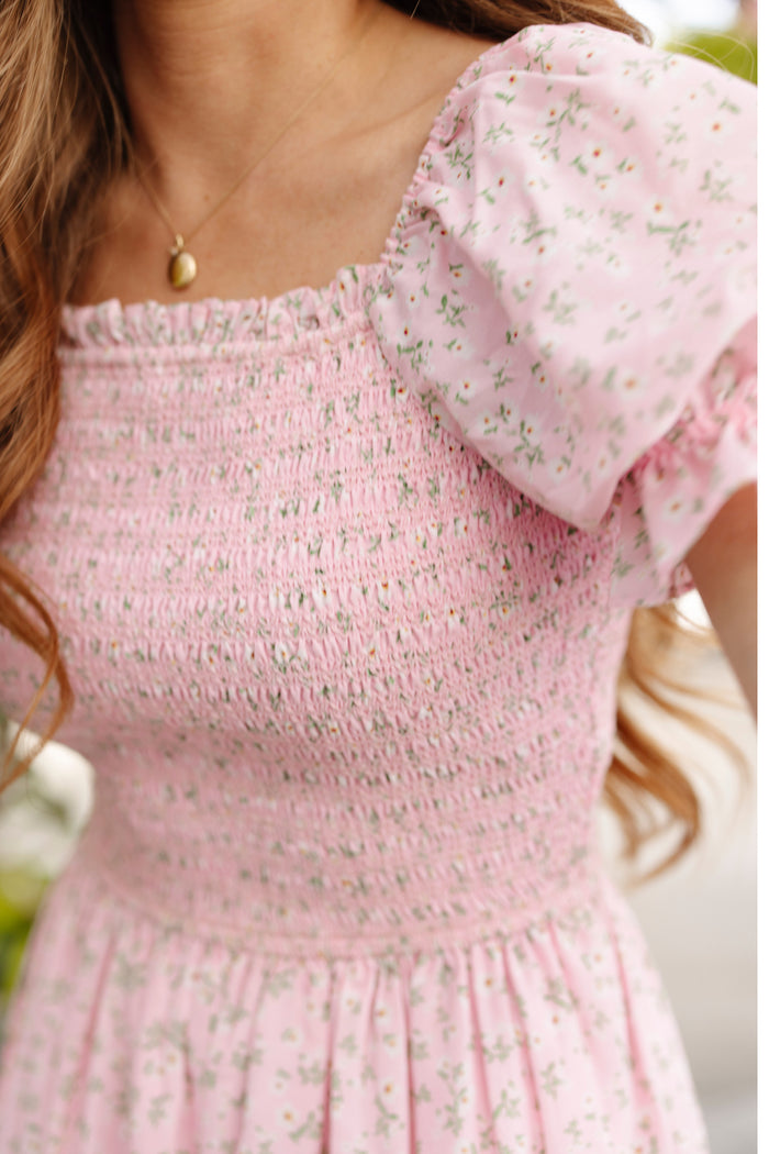 A woman in the Ivy City Co Valencia Dress in Pink Ditsy Floral, a maternity-friendly smocked style with puff sleeves, wears a gold pendant necklace. Her long wavy hair frames her face; the cropped photo highlights her chic upper body look.