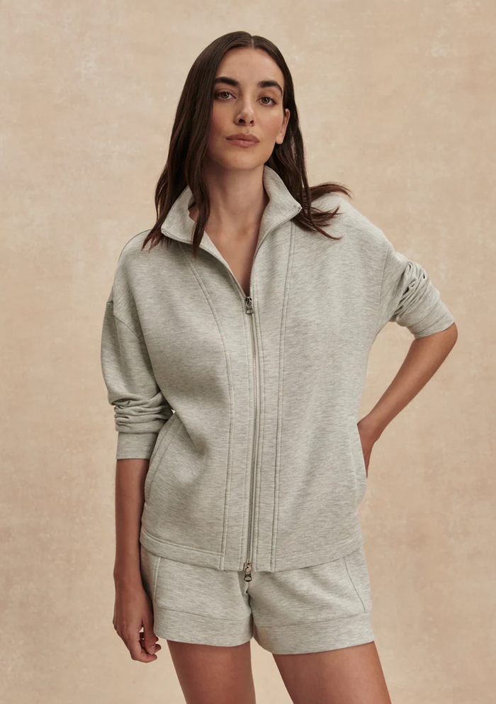 A woman modeling a light heather gray zip-up sweatshirt and matching shorts set against a textured beige studio backdrop.