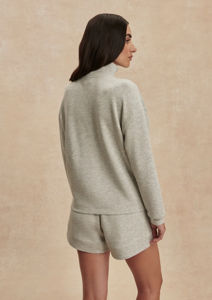 A woman shown from the back wearing a light heather gray mock-neck long-sleeve top and matching shorts against a textured beige studio backdrop.