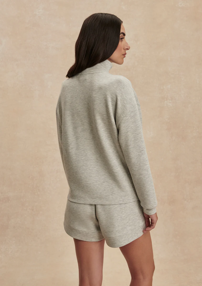 A woman shown from the back wearing a light heather gray mock-neck long-sleeve top and matching shorts against a textured beige studio backdrop.