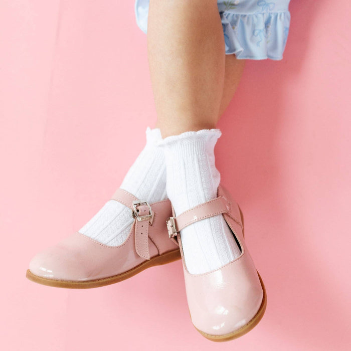 A child wearing Little Stocking Co. White Frill Midi Socks and shiny light pink Mary Jane shoes with buckles sits against a soft pink background, with only their lower legs and feet visible.