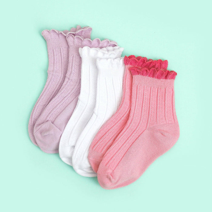 Three pairs of Little Stocking Co.’s White Frill Midi Socks are displayed on a mint green background. These soft cotton socks feature delicate ribbing and charming scalloped tops in pastel hues: lavender, white, and pink.